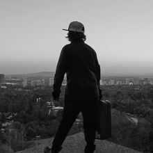 Schwarz-weiß Foto. Ein Mensch, der von hinten zu sehen ist, steht am Rande einer Plattform und hat in der rechten Hand einen kleinen Koffer. Hinter dieser Szene liegen tief unten Wald und Stadt mit Hochhäusern. Der Ausblick ist weit.