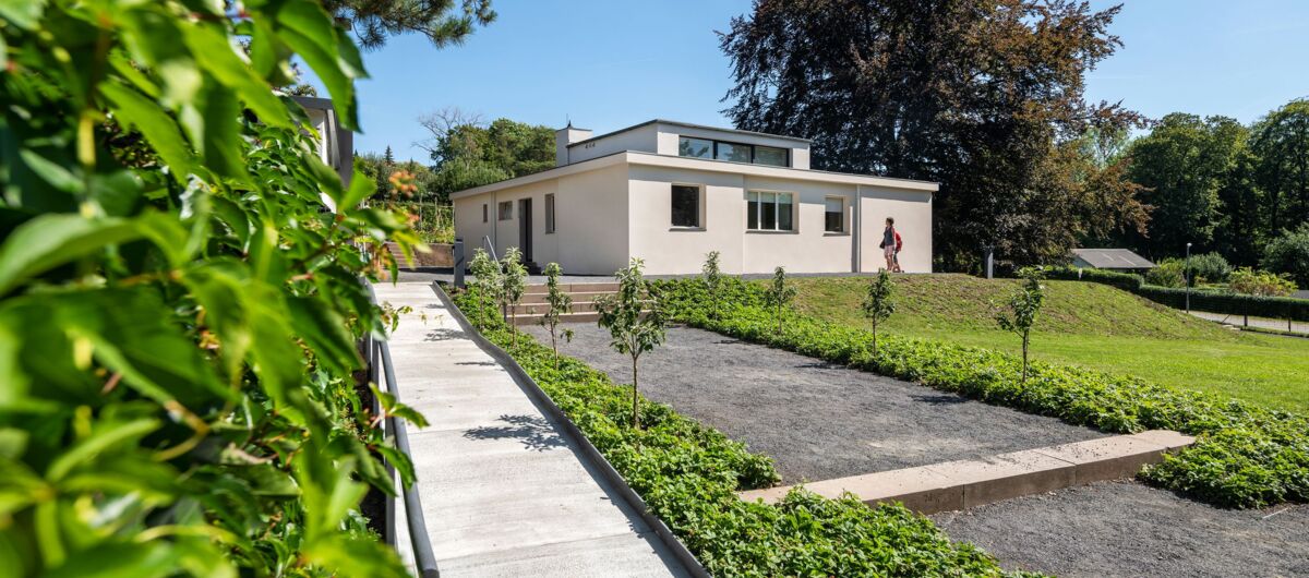 Zweistöckiges Haus mit kleinem Aufbau, weißer Stein. Vereinzelte Fenster in den Wänden. Das Haus steht in einem Garten, welcher von einer breiten Treppe geteilt wird.