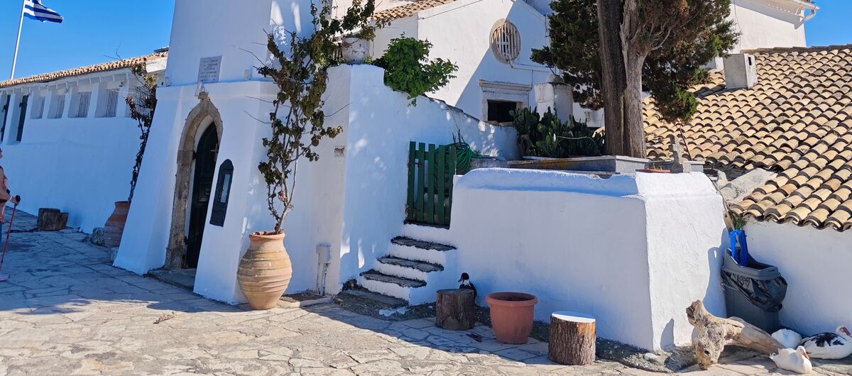 Foto eines weißen Gebäudes im griechischen Stil vor blauem Himmel. Über einer Tür ist ein Turm mit einer Glocke. Vor dem Gebäude ist ein Natursteinboden. Auf dem Gebäude weh eine griechische Flagge.