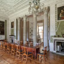 Foto des Speisesaals des Schloss Wörlitz. Kunstvoll verzierte weiße Wände werden durch einen von vier Säulen geschützten Erker sowie durch einen Kamin rechts daneben unterbrochen. Vor den Säulen sind 3 ausklappbare Holztische mit 14 Holzstühlen aufgebaut.