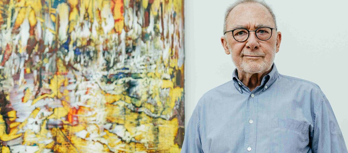 Gerhard Richter mit grauem Haar, Brille und hellblauem Hemd steht vor einem großformatigen, farbenfrohen abstrakten Gemälde. Das Bild im Hintergrund zeigt eine lebendige Mischung aus Gelb-, Rot- und Blautönen.