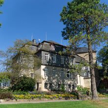 Foto eines viergeschossigen Schlosses, das verschachtelt mit Erkern und Türmchen gebaut wurde. Davor strahlt in bunten Farben ein üppiges Blumenbeet. Um das Haus stehen hohe Bäume. Im Vordergrund ist eine Rasenfläche. Der Himmel ist blau.