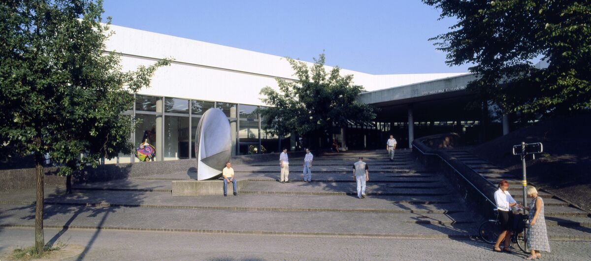 Foto des Eingangs vom Sprengel Museum Hannover. Langezogene flache Treppenstufen führen zu dem flachen Gebäude mit Glasfront und überdachtem Eingang. Eine abstrakte Skulptur steht auf den Stufen. Sieben Menschen halten sich auf den Stufen auf.