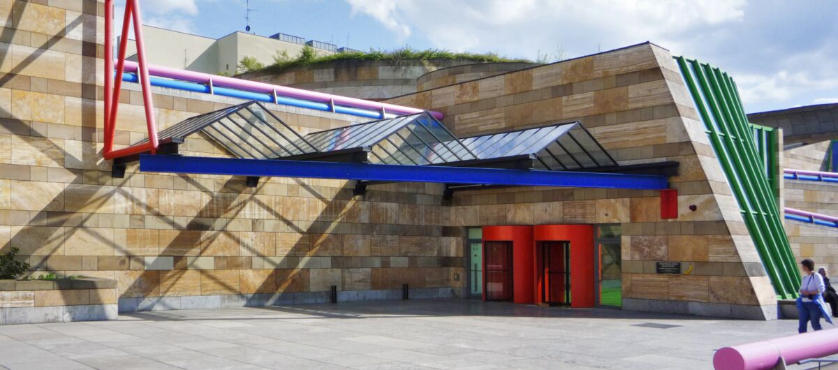 Außenansicht der Staatsgalerie Stuttgart. Die Fassade besteht aus gelben Sandsteinquadern, die durch bunte Metallelemente und einer Glasüberdachung im Zickzackmuster verziert ist.