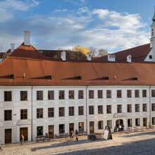 Foto eines breiten dreigeschossigen Hauses mit Ziegelrotem Dach inmitten einer Stadt. In der Breite zieren jeweils 21 Fenster die Front des 1. und 2. Geschosses. Die Front des Erdgeschosses wird ähnlich symmetrisch durch Fenster und Türen geschmückt.