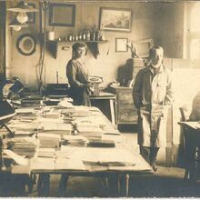 Schwarz-weiß Foto im Querformat eines Handwerksbetriebs. In dem Raum sind ein großer Tisch mit vielen Stapel Papier sowie weitere Arbeitsplätze und Schränke zu sehen. Zwei Frauen schauen direkt in die Kamera. Zwei Männer sind in ihr Tun vertieft.