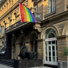 Foto der Außenansicht des Von der Heydt-Museums. Zu sehen ist der Eingangsbereich, der durch zwei abstrakte schwarze Skulpturen gesäumt wird. Auf höhe der ersten Etage hängt eine Fahne des Museums sowie eine Regenbogenfahne.