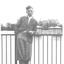 Schwarz-weiß Foto des jungen Willy Brandts in Jackett und Knickerbocker. Er steht vor einem Gewässer und lehnt sich mit seinem linken Arm auf einen Zaun. Die rechte Hand Steckt in seiner Hosentasche. Er schaut mit neutralem Gesichtsausdruck nach links.