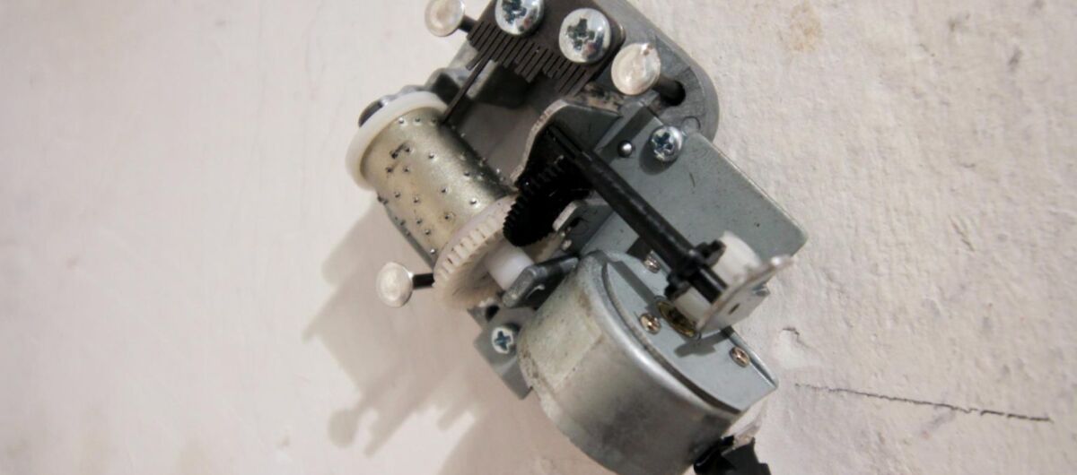 Mechanische Spieluhr ohne Gehäuse, von der der Klangkörper mit seiner Spule und den Punktförmigen Erhebungen zu sehen ist. Die Kurbel ist an ein Stromkabel angeschlossen. Das elektrifizierte Musikinstrument ist mit Schrauben an einer Wand befestigt.
